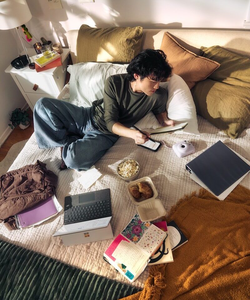 Jonge man ligt op een bed met een laptop en snacks, terwijl hij geconcentreerd naar het scherm kijkt in een informele slaapkameromgeving.