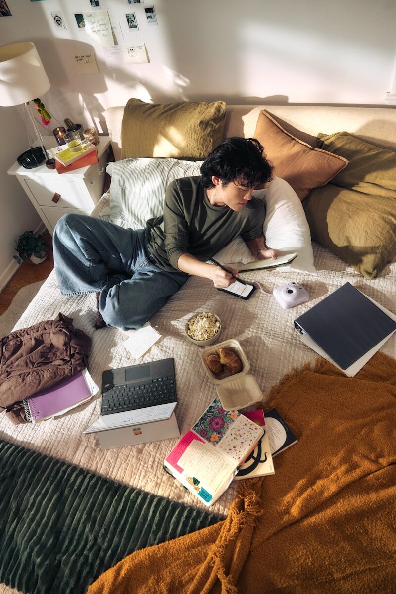 Jonge man ligt op een bed met een laptop en snacks, terwijl hij geconcentreerd naar het scherm kijkt in een informele slaapkameromgeving.