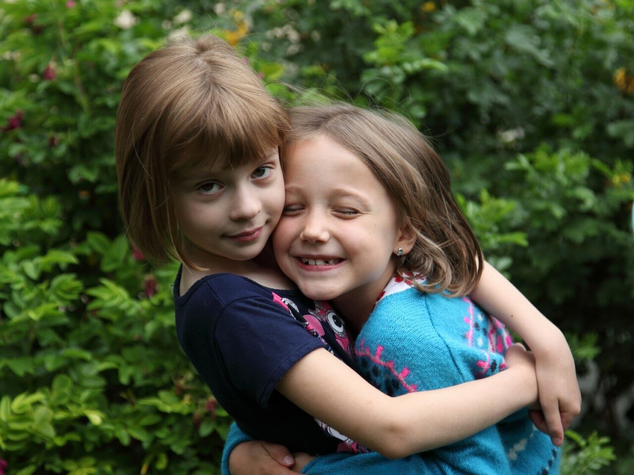 Two young girls hugging outdoors with greenery behind them