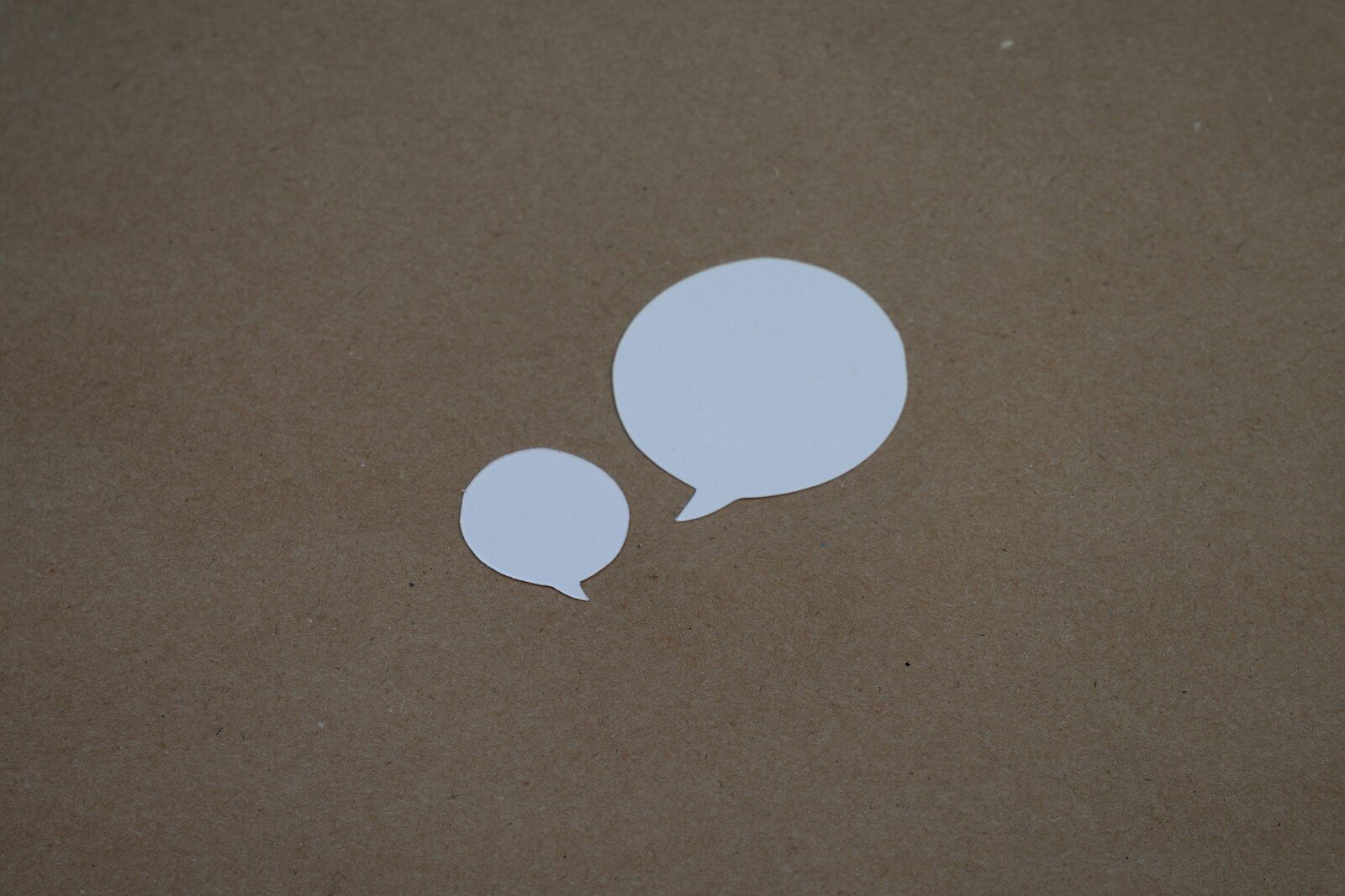 Twee witte tekstballonnen, een grote en een kleine, liggen op een bruine achtergrond. De afbeelding symboliseert communicatie, gesprekken en de ontmoeting tussen verschillende perspectieven.