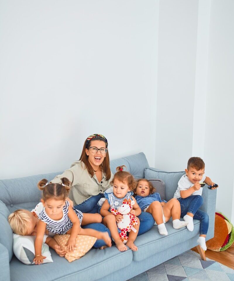 Vrouw zit op een bank samen met vijf jonge kinderen in een huiselijke omgeving, als illustratie van kinderopvang, opvoeding en gezinsleven.